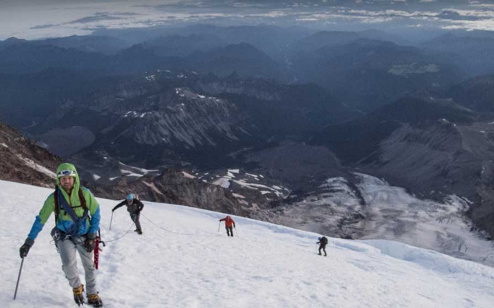 High above a cast mountainous landscape, four people wearing safety gear make their way up a snowy incline.